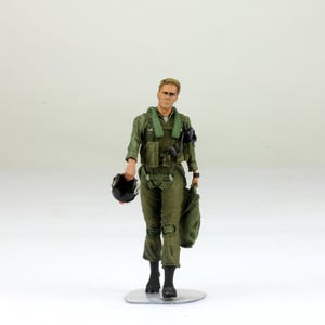 Puede incluir: Figura en miniatura de un piloto con un traje de vuelo verde, sosteniendo un casco negro y una bolsa de lona verde. El piloto tiene el pelo rubio corto y está de pie sobre una base circular transparente. La figura es detallada.