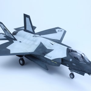 May include: A model of a gray and white fighter jet with a clear cockpit. The jet has a black and white camouflage pattern and the text "US AIR FORCE 155021" on the tail.