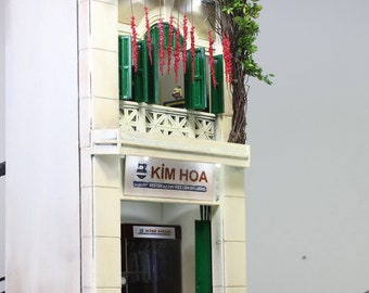 May include: A miniature model of a two-story building with a green awning and a sign that reads "KIM HOA" in red letters. The building has a white facade with red decorations hanging from the top floor. The model is made of wood and plastic.
