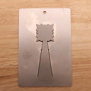 May include: A silver metal card with a decorative cutout design. The card has a rectangular shape with a small hole at the top and a floral-like design in the center. The card is placed on a wooden surface.