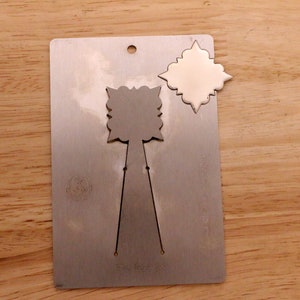 May include: A silver metal die-cut card with a decorative design. The card has a hole at the top and a cut-out shape of a stylized flower. A separate, identical shape is detached from the card.