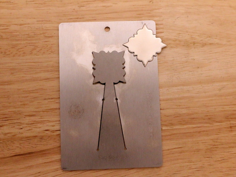 May include: A silver metal die-cut card with a decorative design. The card has a hole at the top and a cut-out shape of a stylized flower. A separate, identical shape is detached from the card.