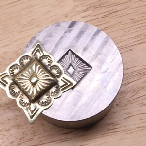 Puede incluir: Primer plano de una herramienta de estampado de metal y una pieza decorativa de metal. La pieza met&aacute;lica tiene un dise&ntilde;o cuadrado central con l&iacute;neas radiales y un borde floral. La herramienta es un bloque de metal circular plateado con un dise&ntilde;o estampado a juego.