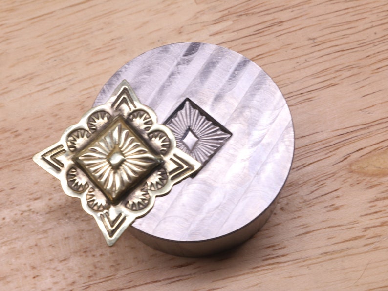Puede incluir: Primer plano de una herramienta de estampado de metal y una pieza decorativa de metal. La pieza met&aacute;lica tiene un dise&ntilde;o cuadrado central con l&iacute;neas radiales y un borde floral. La herramienta es un bloque de metal circular plateado con un dise&ntilde;o estampado a juego.