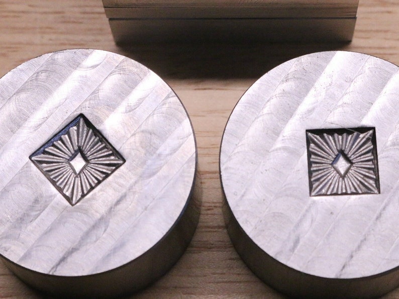 Puede incluir: Dos sellos met&aacute;licos circulares con un dise&ntilde;o en forma de diamante en el centro. El dise&ntilde;o presenta un diamante m&aacute;s peque&ntilde;o rodeado de l&iacute;neas radiales. El metal tiene un acabado cepillado, y los sellos probablemente se utilizan para imprimir o grabar en relieve.