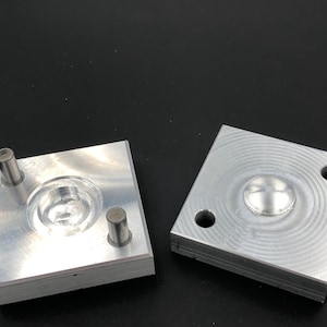 Może przedstawiać: Dwa obrobione bloki aluminiowe z okrągłymi i półkulistymi wgłębieniami. Jeden blok ma dwa cylindryczne kołki. Bloki mają szczotkowane metalowe wykończenie i leżą na czarnej powierzchni. Prawdopodobnie używane do produkcji lub formowania.