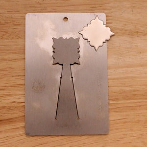 May include: A silver metal die-cut template with a decorative design. The template has a rectangular shape with a hole at the top and a cut-out design in the center. A separate, matching die-cut shape is next to the template.