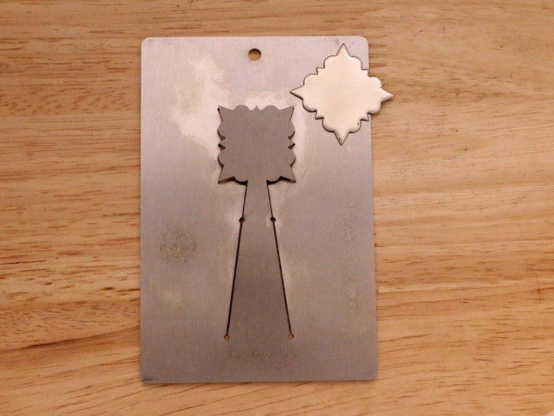 May include: A silver metal die-cut template with a decorative design. The template has a rectangular shape with a hole at the top and a cut-out design in the center. A separate, matching die-cut shape is next to the template.