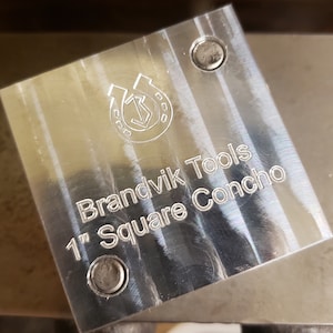 Puede incluir: Una herramienta met&aacute;lica cuadrada y pulida con el texto "Brandvik Tools 1" Square Concho." La herramienta tiene un logotipo de herradura y flecha y dos peque&ntilde;os agujeros. El cuadrado mide 2,54 cm.