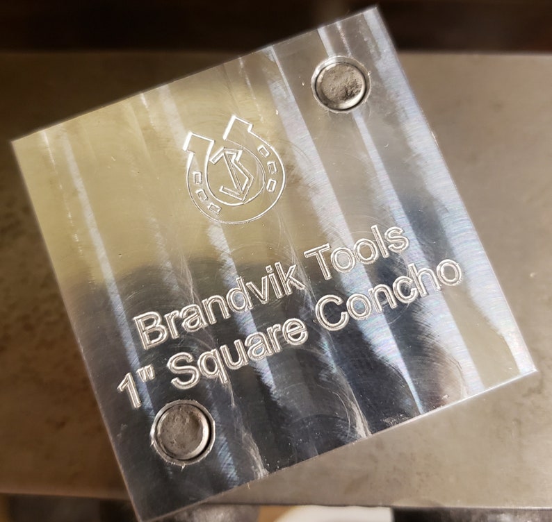 Puede incluir: Una herramienta met&aacute;lica cuadrada y pulida con el texto "Brandvik Tools 1" Square Concho." La herramienta tiene un logotipo de herradura y flecha y dos peque&ntilde;os agujeros. El cuadrado mide 2,54 cm.