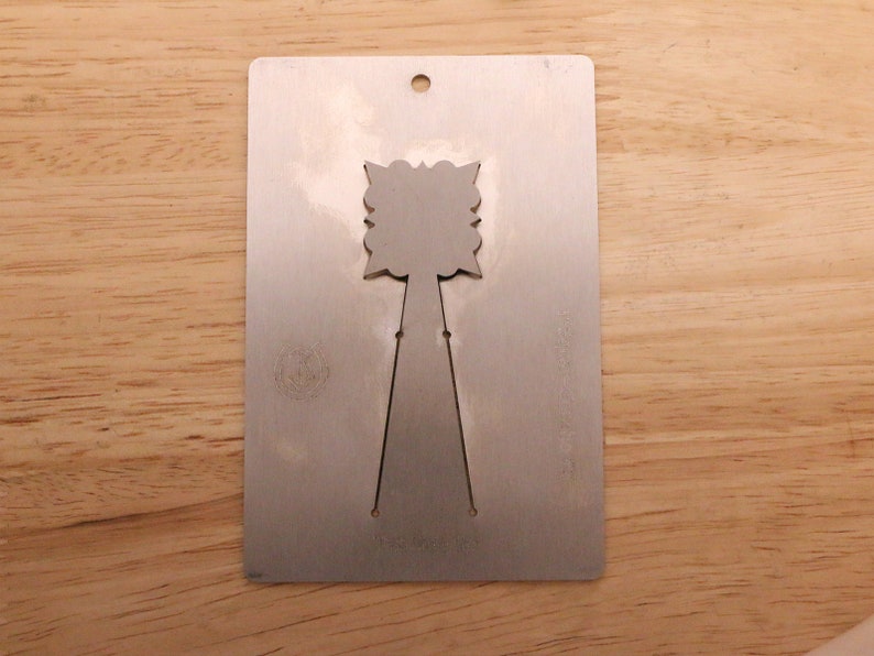 May include: A silver metal card with a decorative cutout design. The card has a rectangular shape with a small hole at the top and a floral-like design in the center. The card is placed on a wooden surface.