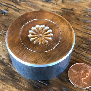 Op de afbeelding: Een ronde, bruine container met een zilveren bloemmotief op het deksel. De container heeft een donkergrijze basis. De container heeft een diameter van ongeveer 3,8 cm. Er ligt een munt naast.