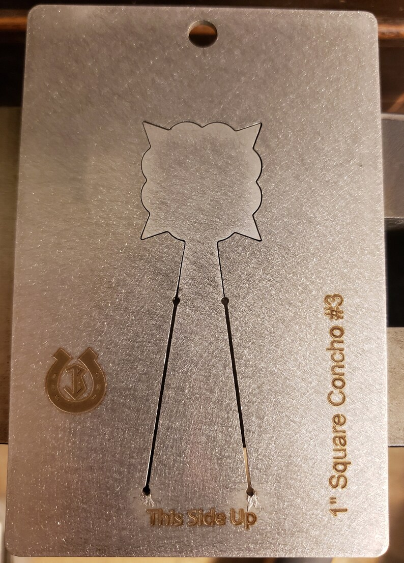 Puede incluir: Una tarjeta de concho de metal plateado con un dise&ntilde;o recortado. La tarjeta tiene un agujero en la parte superior y el texto "1" Square Concho #3" y "This Side Up". Tambi&eacute;n es visible un logotipo de herradura.