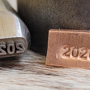 May include: Close-up of metal stamping tools. One tool is stamped with the word "asos" in lowercase letters. A small copper tag is stamped with the number "2026". The tools are on a textured surface.