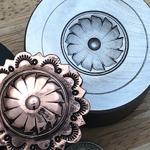 Op de afbeelding: Close-up van metaalbewerkingstools, waaronder een koperkleurig, decoratief metalen stuk met een bloemenontwerp en een bijpassende matrijs. De matrijs is zilverkleurig en heeft een vergelijkbaar bloemenpatroon. De tools liggen op een houten oppervlak.