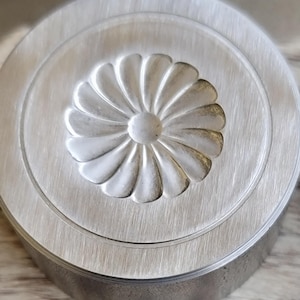 Op de afbeelding: Een cirkelvormig, geborsteld metalen object met een verhoogd bloemmotief in het midden. De bloem heeft meerdere gebogen bloemblaadjes die uitstralen vanuit een centraal punt. Het object heeft een afgeronde rand en een getextureerd oppervlak.