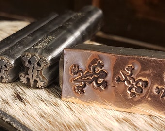 May include: Close-up of metal tools for stamping a lizard design. Three metal stamps with lizard shapes are next to a copper plate with stamped lizard impressions. The tools are on a textured, light brown surface.