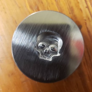 Puede incluir: Objeto metálico circular plateado con acabado cepillado. El centro presenta un diseño de calavera en relieve. Probablemente es un elemento decorativo, con un diámetro de aproximadamente 5 cm.