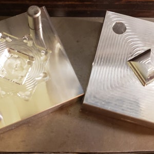 Puede incluir: Dos moldes de metal plateado, cada uno cuadrado con un dise&ntilde;o geom&eacute;trico. Un molde tiene una cavidad en forma de estrella con un cuadrado central, y el otro tiene forma de diamante en relieve. Los moldes est&aacute;n sobre una superficie met&aacute;lica.