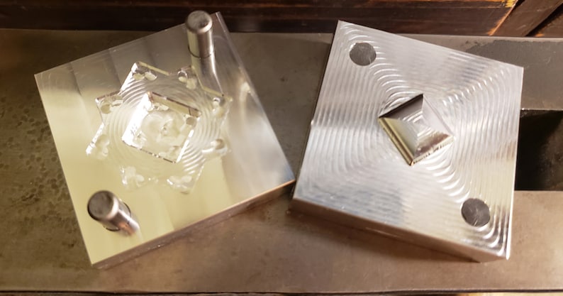 Puede incluir: Dos moldes de metal plateado, cada uno cuadrado con un dise&ntilde;o geom&eacute;trico. Un molde tiene una cavidad en forma de estrella con un cuadrado central, y el otro tiene forma de diamante en relieve. Los moldes est&aacute;n sobre una superficie met&aacute;lica.