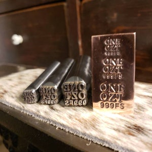May include: A set of metal stamps and a small bar. The stamps are labeled "ONE OZ" and the bar is marked "ONE OZ 999FS". The items are displayed on a textured surface.