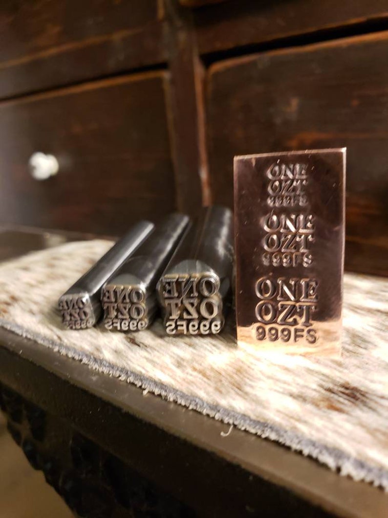May include: A set of metal stamps and a small bar. The stamps are labeled "ONE OZ" and the bar is marked "ONE OZ 999FS". The items are displayed on a textured surface.