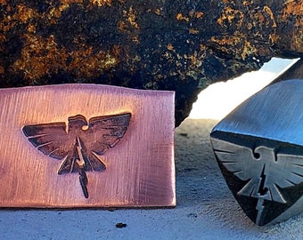 May include: A copper metal stamp with a rectangular shape and an embossed eagle design. Next to it is a metal tool with a similar eagle design. The eagle has outstretched wings and a lightning bolt.