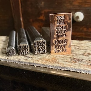 May include: A copper-colored metal bar and metal stamps with the text "ONE OZT 999FS" are displayed. The bar is rectangular with the text repeated three times. Three stamps are in the foreground, each with the same text in reverse.