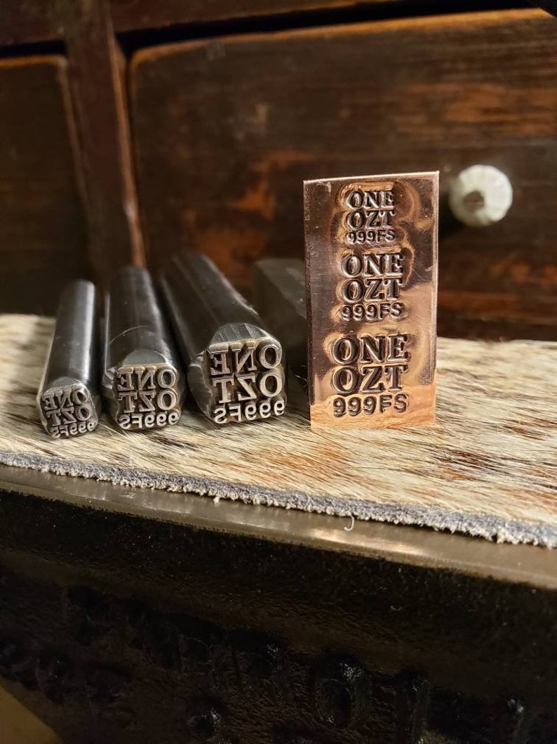 May include: A copper-colored metal bar and metal stamps with the text "ONE OZT 999FS" are displayed. The bar is rectangular with the text repeated three times. Three stamps are in the foreground, each with the same text in reverse.