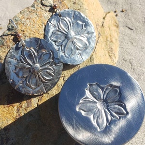 May include: Silver-coloured, hammered metal earrings and a matching pendant with a floral design. The earrings have a hook closure and a small copper-coloured bead. The pendant is a flat, circular shape with a raised flower design.