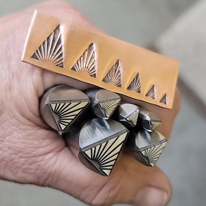 Puede incluir: Un conjunto de sellos metálicos con un diseño de sol. Los sellos se sostienen en una mano e incluyen una pieza rectangular de cobre y varios sellos en forma de diamante. El diseño del sol está grabado en el metal.