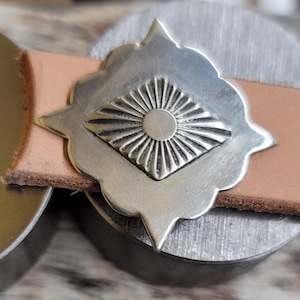 Op de afbeelding: Close-up van een zilveren metalen decoratieve versiering met een sunburst-ontwerp, geplaatst op een lichtbruine leren riem. De versiering heeft een stervorm met een centraal sunburst-patroon.
