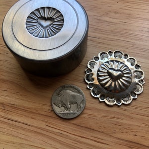 Puede incluir: Una herramienta de estampado de metal plateado y una concha decorativa de metal plateado con un diseño de corazón. Una moneda estadounidense de cinco centavos está al lado de la concha. La herramienta de estampado es cilíndrica con un diseño de corazón en el centro.