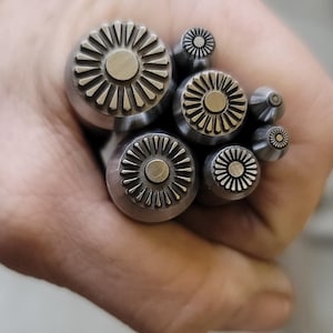 May include: A collection of metal stamps with a sunburst design. The stamps vary in size and are held in a hand. The stamps have a circular shape with a central dot and radiating lines.