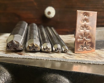 May include: A set of metal stamping tools featuring floral patterns. The tools are of varying sizes and shapes, with detailed floral designs on the striking surfaces. A copper-coloured metal piece shows stamped floral impressions. The tools are arranged on a dark surface.