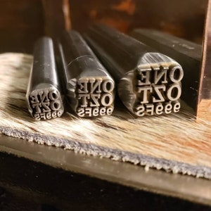 May include: Close-up of metal stamps with the word "ONE" and the number "2" in a serif font. The stamps are arranged on a wooden surface with a textured, light brown background. The stamps are used for leathercraft or similar projects.