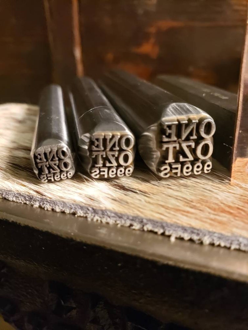 May include: Close-up of metal stamps with the word "ONE" and the number "2" in a serif font. The stamps are arranged on a wooden surface with a textured, light brown background. The stamps are used for leathercraft or similar projects.