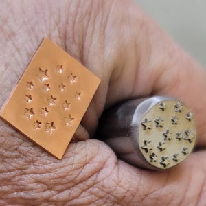 May include: A copper metal stamp and a metal punch tool, both featuring a star pattern. The stamp is rectangular, and the punch is cylindrical with a circular star design. The tools are used for metalworking and crafting.