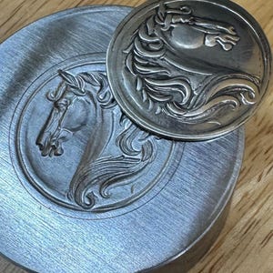 Puede incluir: Un sello y una moneda de metal plateado con un relieve detallado de la cabeza y la melena de un caballo. La moneda descansa sobre el sello, sobre una superficie de madera.