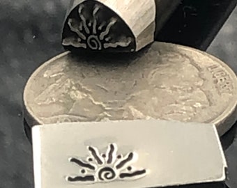May include: A metal stamping tool with a sunburst design, alongside a stamped impression and a coin. The sunburst design features radiating lines and a central spiral. The tool is used for metalworking and crafting.