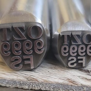 May include: Close-up of two metal stamps with the letters "TSO", followed by "eeee" and the number "27". The stamps are silver and have a rounded top. The text is in a serif font.