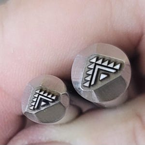 Puede incluir: Dos sellos metálicos con un diseño geométrico. Los sellos son redondos con un borde facetado y presentan un patrón angular blanco y negro. Los sellos probablemente se utilizan para manualidades o joyería.