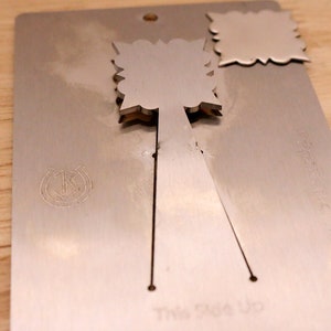 May include: A silver-colored metal card with a decorative cutout design. The card has a horseshoe and anchor emblem and the words "This Side Up" printed on it. The card is on a wooden surface.