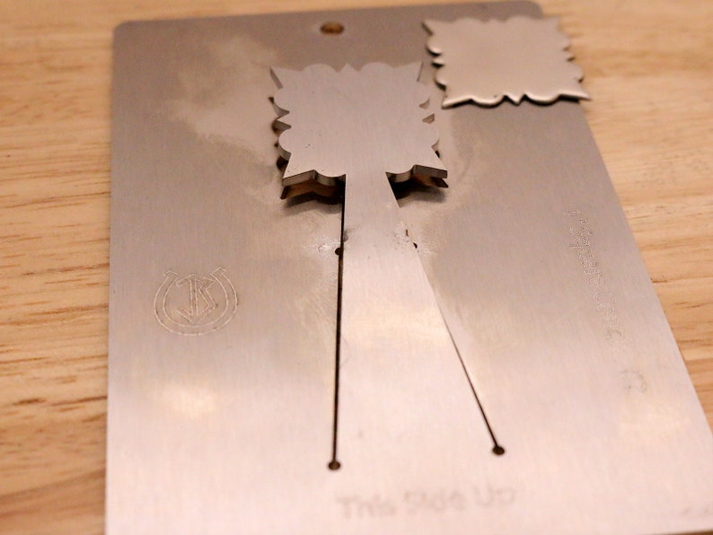 May include: A silver-colored metal card with a decorative cutout design. The card has a horseshoe and anchor emblem and the words "This Side Up" printed on it. The card is on a wooden surface.