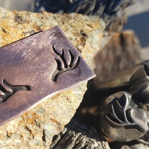 May include: A rectangular, copper-colored metal stamp with a stylized antler design, alongside two matching metal rings. The items are displayed on a textured rock surface, with natural light enhancing the details.
