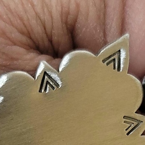 May include: Close-up of a silver-colored metal pendant with a floral design and geometric accents. The pendant is next to a small, geometric-shaped metal piece. The design features etched arrow-like shapes.