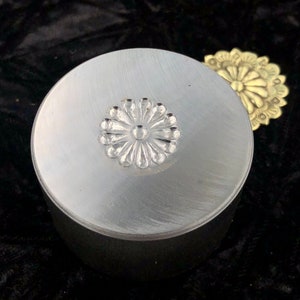 Op de afbeelding: Een zilverkleurige, ronde container met een bloemenontwerp op het deksel. Het deksel heeft een geborstelde metalen afwerking en een verhoogd bloemmotief in het midden. Een goudkleurig bloemenaccent is zichtbaar op de achtergrond.