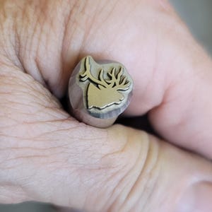 May include: A metal stamp featuring a deer head design. The stamp has a gold-coloured silhouette of a deer's head and antlers, set against a silver base. The stamp is held in a hand.