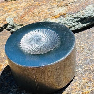 May include: A silver-colored, cylindrical object with a floral design on the top surface. The object is sitting on a rough, brown and gray stone surface. The floral design is in the center of the top surface.