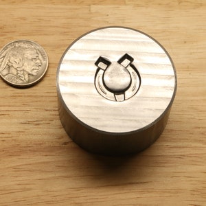 May include: A silver-colored metal stamp with a horseshoe design is shown next to a nickel coin. The stamp is cylindrical with a horseshoe shape carved into the top. The coin is a U.S. nickel.